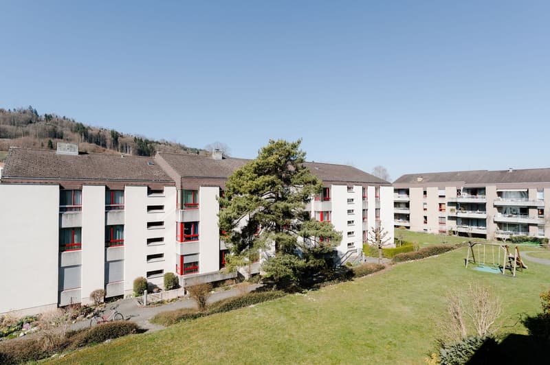 A multi-story apartment building with balconies, red window frames, and a playground in the front yard.