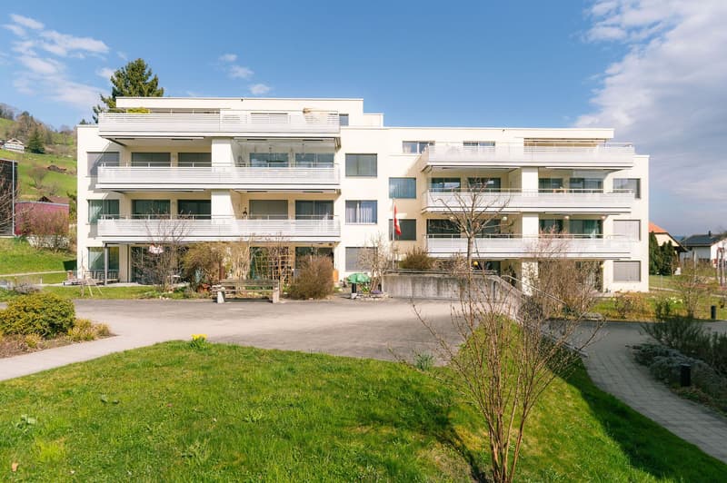 Modern white apartment building with multiple balconies, located on a hill with a view of the surrounding landscape