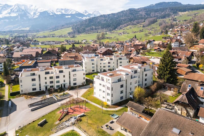 Aerial view of a residential area with several modern white buildings, surrounded by green fields and mountains in the background.