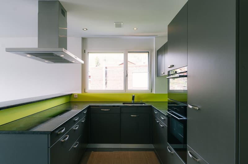 modern kitchen with black countertops, green accent wall, stainless steel range hood, built-in oven and dishwasher, black cabinets, wooden floor