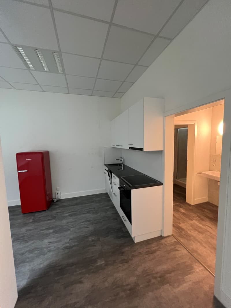 The image shows a studio apartment with a kitchenette area. The kitchenette has a sink, cabinets, and a dishwasher. The floor is a gray laminate or tile material. The ceiling has a grid of recessed lighting panels. There is a red retro-style refrigerator