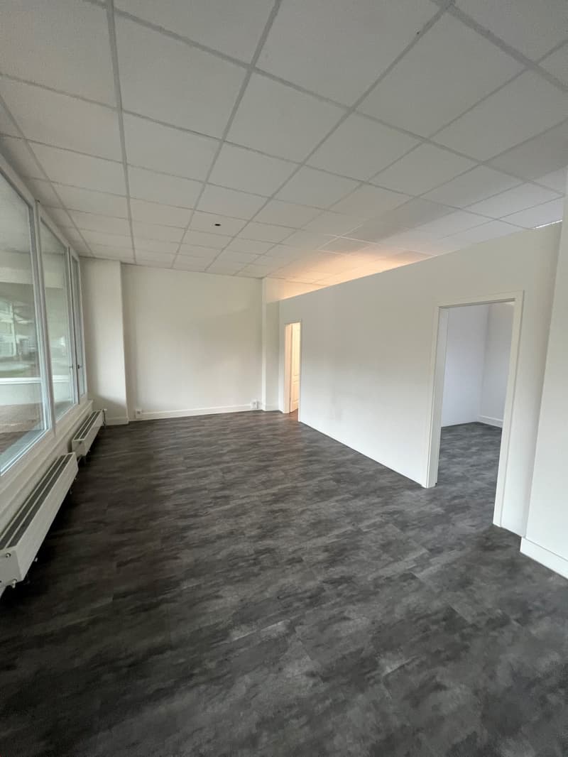 The image shows a large, open office space with a suspended ceiling grid and gray carpeted flooring. The walls are painted a light color, and there are several windows along one side of the room. The space appears to be ready for occupancy, with no furnit