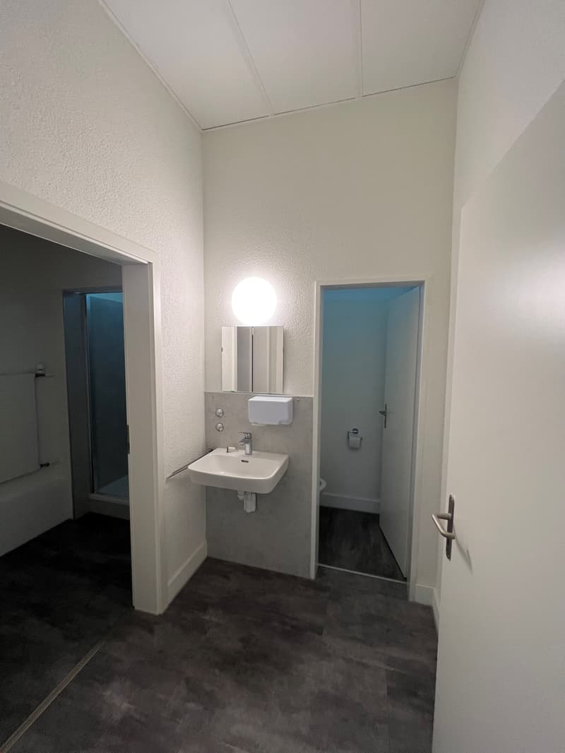 The image shows a bathroom with a white sink, a mirror, and a door leading to another room. The walls are painted in a light color, and the floor is covered with dark tiles.