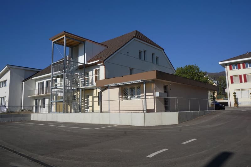 2-story building with a balcony, gray exterior walls, and a tiled roof. There is a staircase leading up to the balcony on the side of the building. The building appears to be located in a residential or commercial area with other buildings nearby.