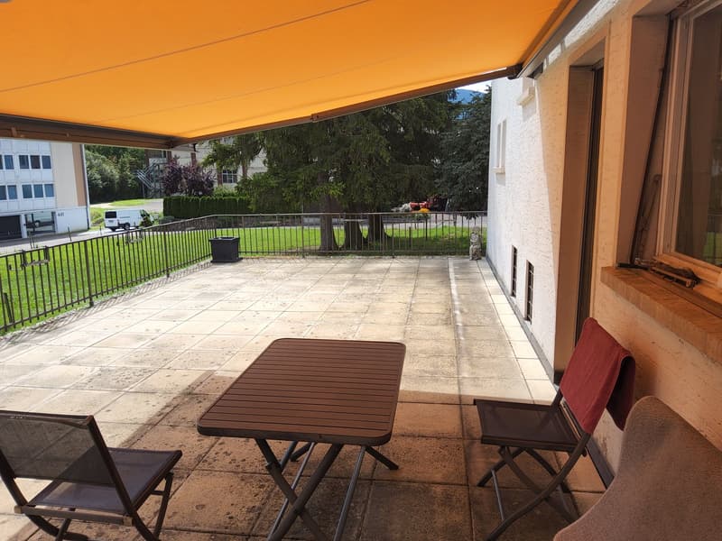 Balcony with yellow awning, black metal railing, square tile floor, a table with two chairs, a potted plant, a view of grass and trees, and a view of a building and vehicles.