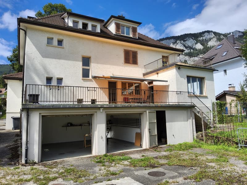 2-story house with a garage and balcony, white walls, brown roof, and fenced garden. Balconies on the second floor with a staircase leading down to the garage.