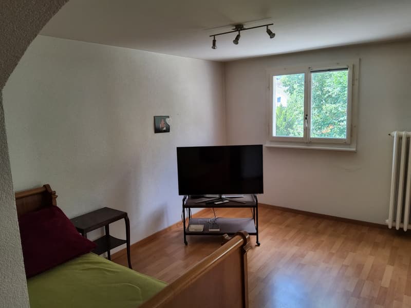 room with wooden floor, white walls, a bed with pillows, a small side table, a flat screen television with a stand, and a window with a view of trees outside