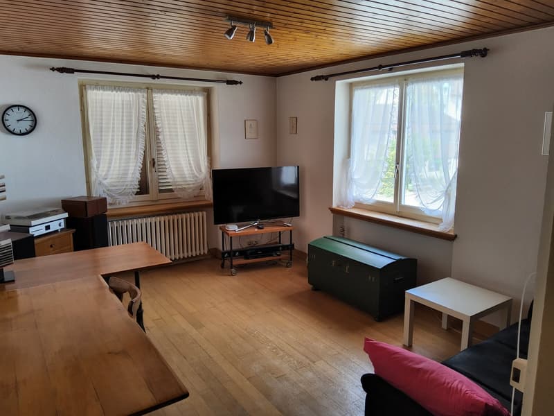 wooden floor, beige walls, two windows with white curtains, large flat screen TV on table, white table, wooden dining table, radiator, clock on wall