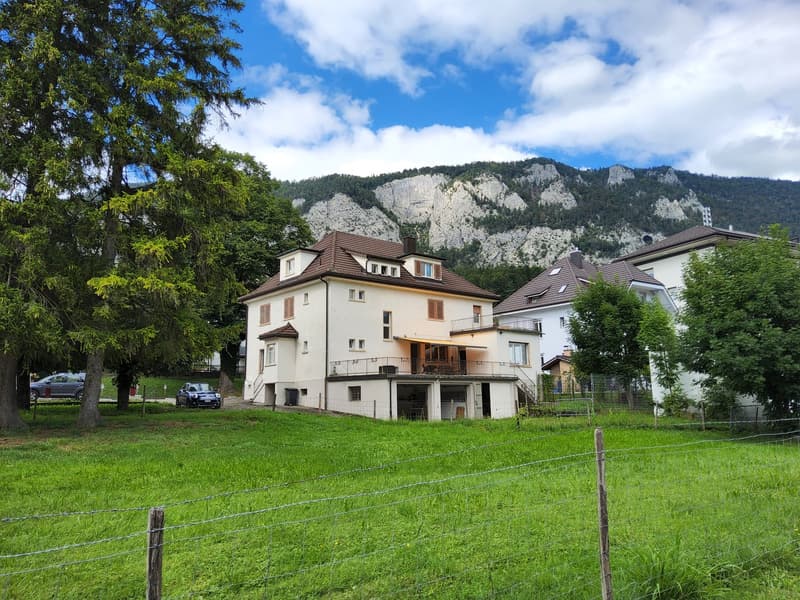 Three-story house, white exterior, brown roof, two neighboring houses, balcony with awning, surrounded by trees, parked cars, green field, barbed wire fence, mountain in the background.