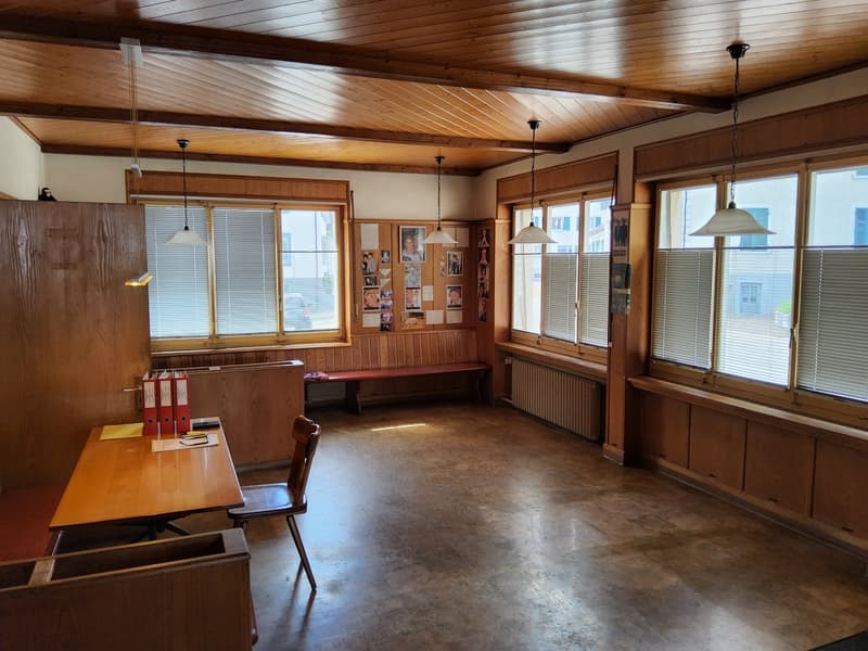 Empty room with wooden flooring, wooden walls, four large windows, hanging lamps, radiators, wooden table, and chair.