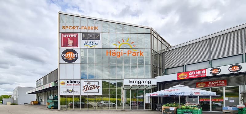 Large glass-fronted building with various retail stores and businesses, including a sports store, a bistro, and a dairy/meat shop. The building has a prominent 'Hagi-Park' sign and an 'Outlet Migros' logo, indicating it is a shopping center or outlet mall