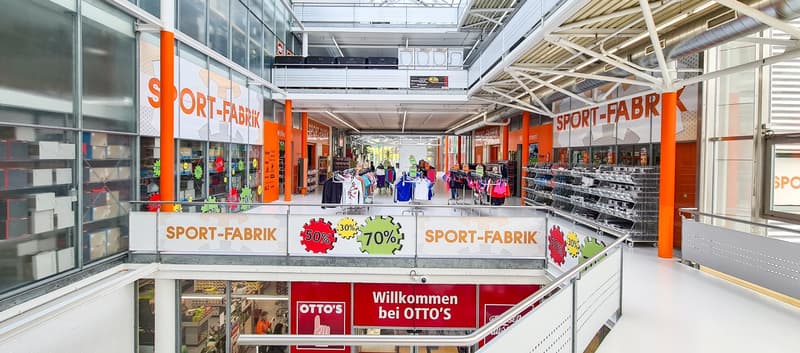 Large indoor sports and athletic goods store with 'Sport-Fabrik' branding, displaying various sports apparel and equipment. The store has a spacious layout with high ceilings, glass walls, and shelves stocked with merchandise. There are signs advertising