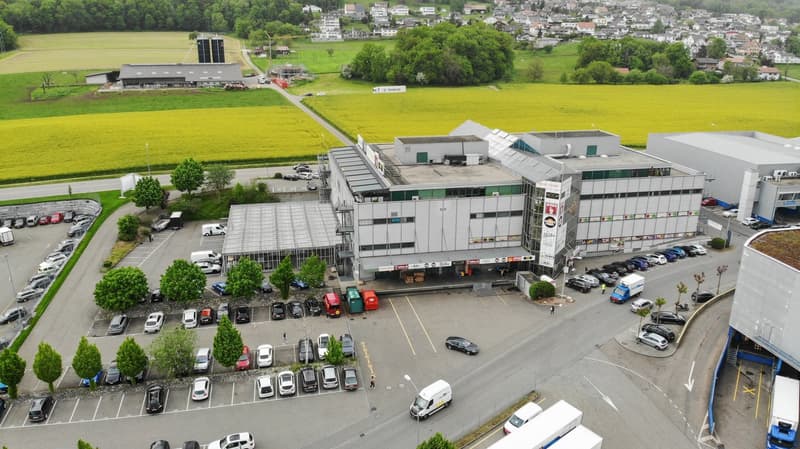 Large commercial building with a parking lot, surrounded by green fields and a small town in the background