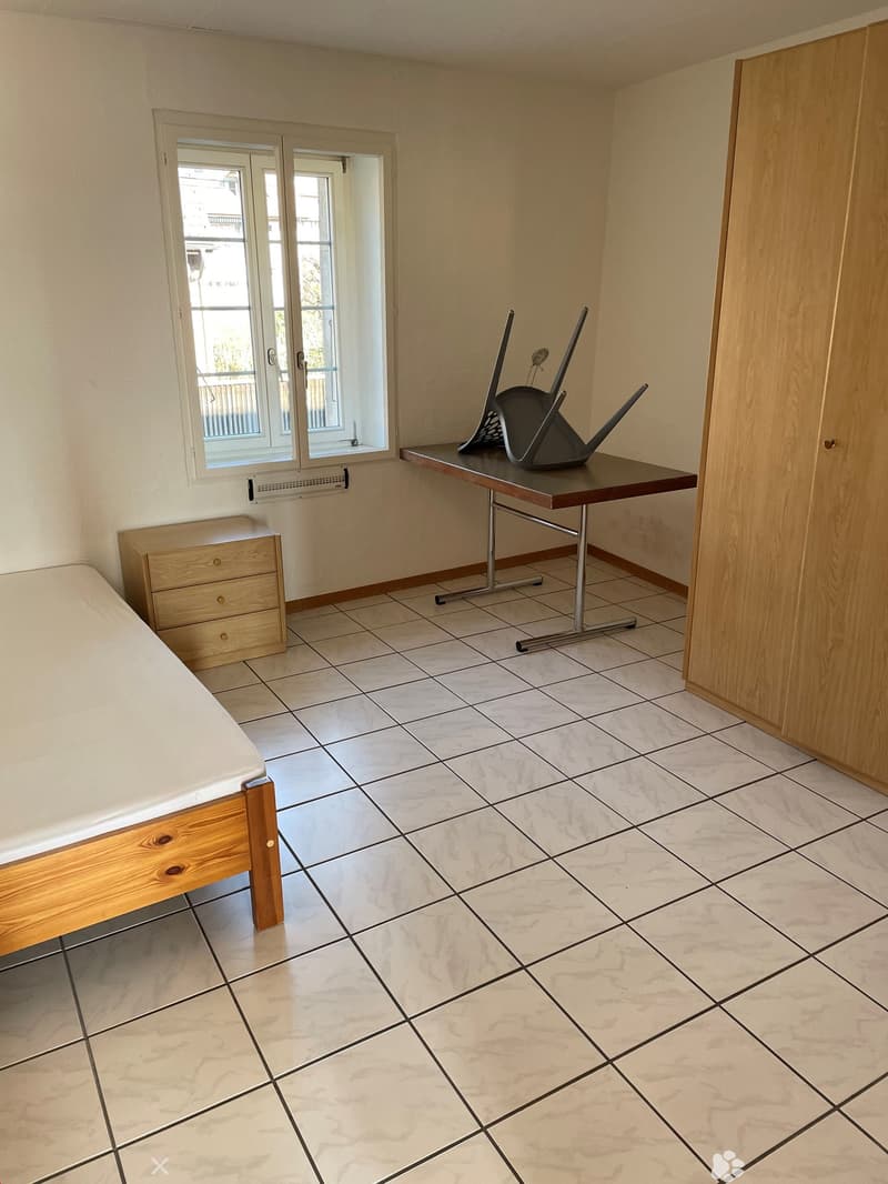 A room featuring white tiles, bed, wooden cabinet, desk with chairs and window