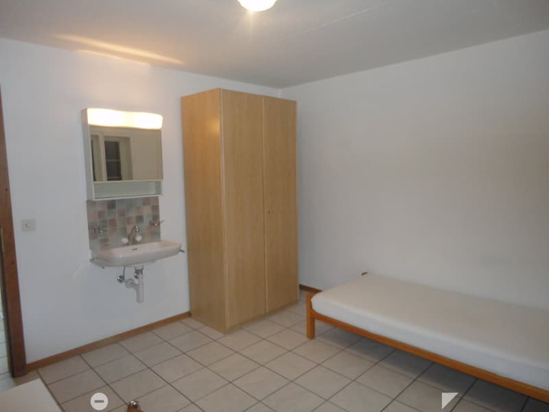 Small room with white tiled floor, white single bed, wooden wardrobe, sink with mirror, power outlet