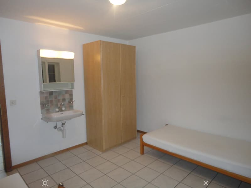Small room with white tiled floor, white single bed, wooden wardrobe, sink with mirror, power outlet