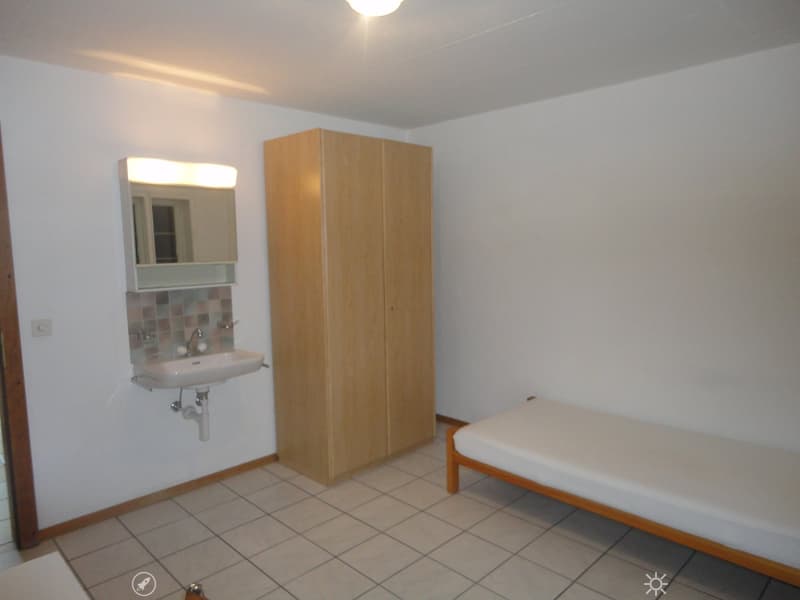 Small room with white tiled floor, white single bed, wooden wardrobe, sink with mirror, power outlet