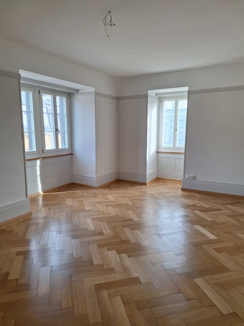 Empty room with wooden parquet flooring, white walls, two windows, power outlets