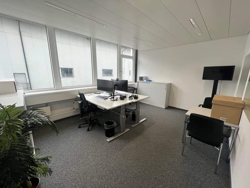 An empty office with carpet floor, large windows, desks, computer monitors, keyboards, chairs, storage boxes, and a television.