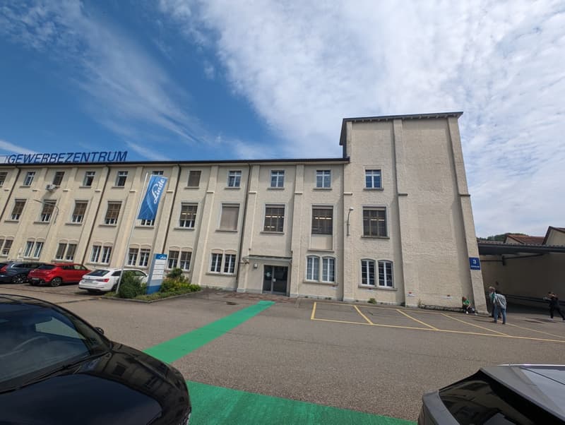 Multi-story office building, multiple parking spaces, large windows, sign reading 'Gewerbezentrum'.