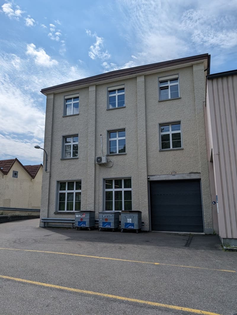 multi-unit building, gray walls, 3 windows per floor, 2nd floor has a balcony, 2 grey garbage bins, garage entrance, street lamp