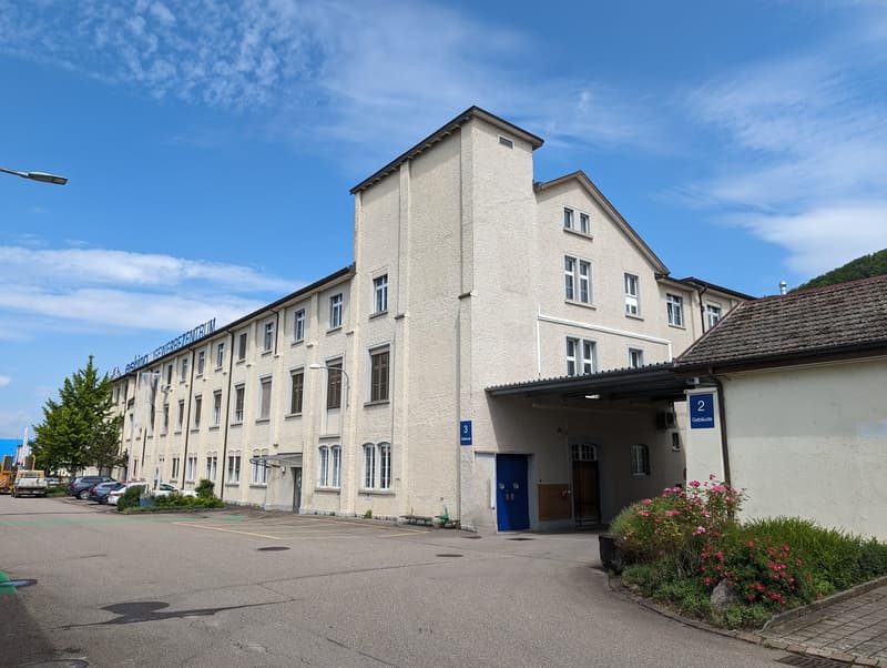 Three-story white building, multiple windows, large parking area in front, blue door, bench, flowering plants.