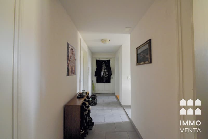 Empty hallway with white walls, tiled floor, wooden shoe rack, picture frames, and a white door.