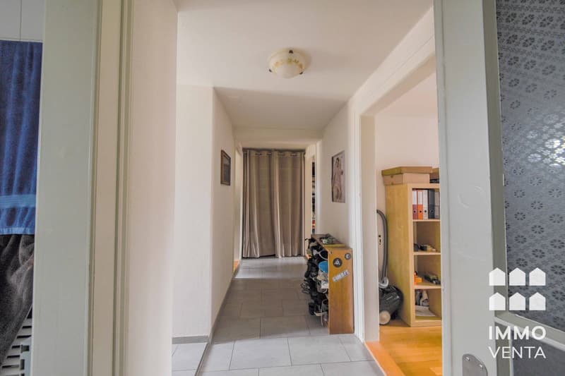 Empty hallway with tiled floor, white walls, ceiling light, wooden shoe rack, and a wooden shelf with books and items.