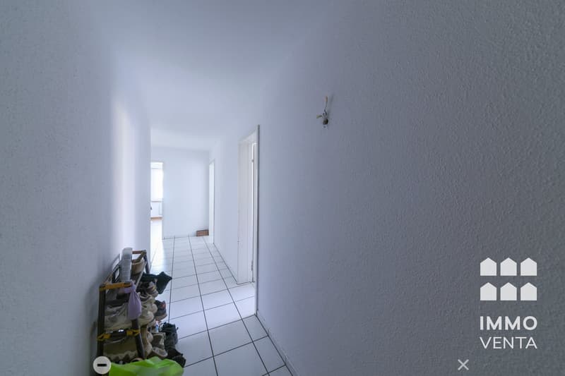 Empty hallway with white tiles, white walls, shoe rack, and a door at the end.