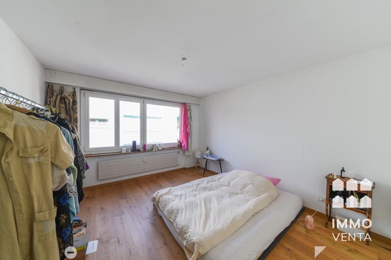 Empty room with wooden floor, white walls, large window, bed, small table, clothes hanging on a rack
