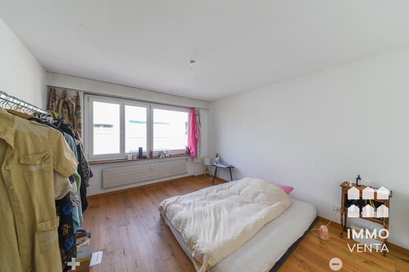 Empty room with wooden floor, white walls, large window, bed, small table, clothes hanging on a rack