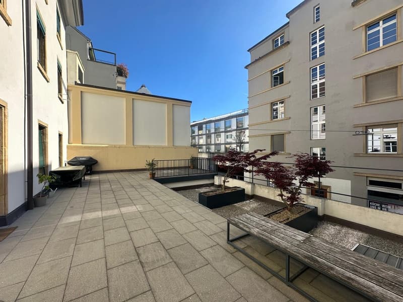 Outdoor area with wooden benches, plants, and a paved floor.