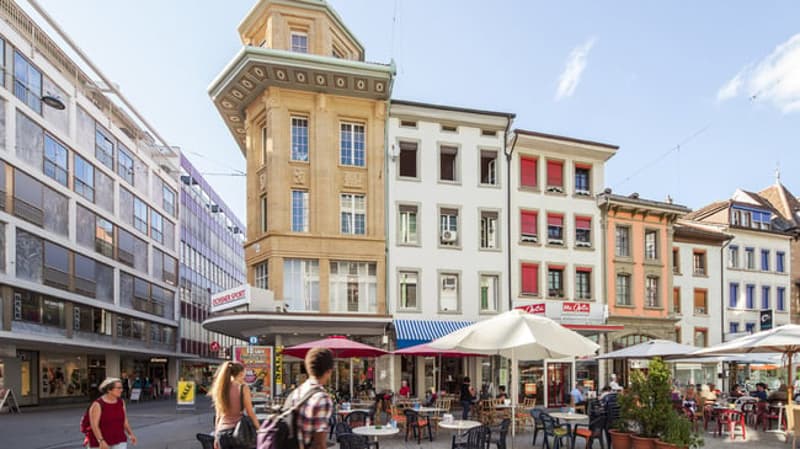 A city street with several buildings, one of which has a tower and a clock, and a few people walking on the street with umbrellas and tables and chairs in front of the buildings.