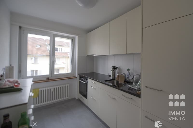modern kitchen with white cabinets, black countertops, white tiled floor, large window, white radiator, built-in oven, sink, and faucet
