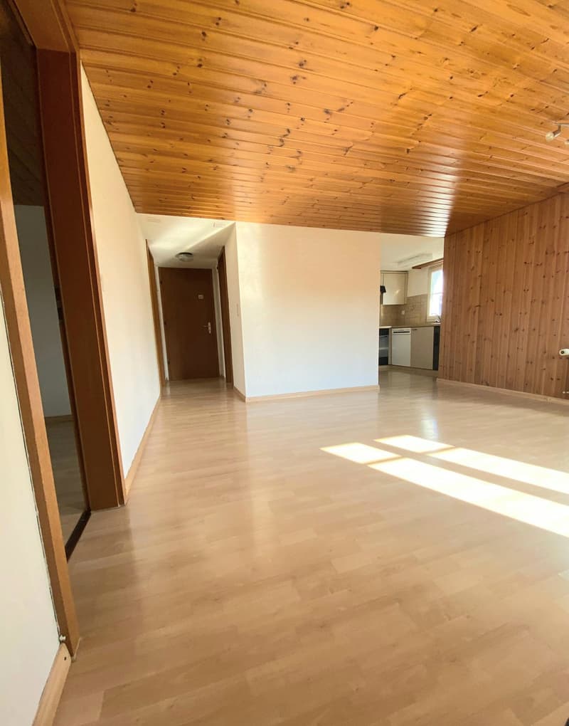 The image shows a spacious open-plan room with a wooden ceiling and hardwood flooring. The room has a clean, minimalist design with white walls and a visible entryway leading to another area, possibly a kitchen. The room appears to be well-lit, with natur