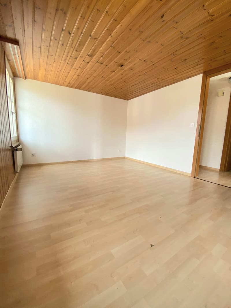 The image shows a room with a wooden ceiling and hardwood floors. The walls are painted white, and there is a door visible in the corner. The room appears to be empty, with no furniture or other objects present.