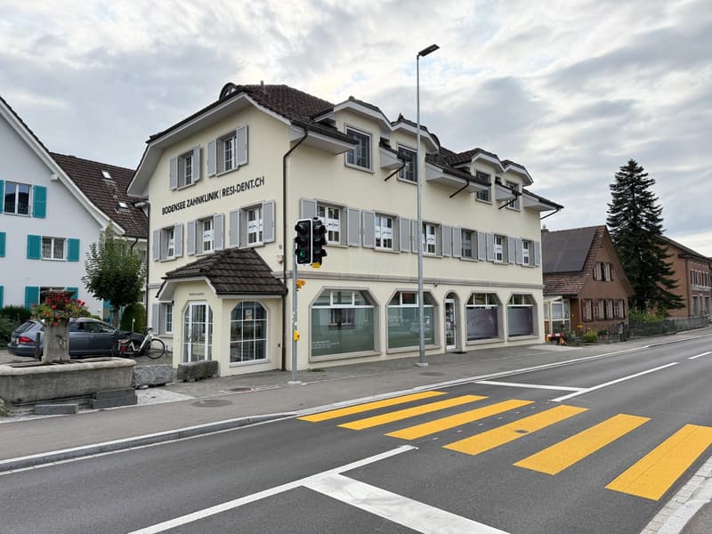 A multi-story beige building with shutters and a brown roof. There is a large car parked in front of the building. The sidewalk in front has a flowerpot, a bench, and a bicycle leaning against the building. Across the street, there is a wide road with a c