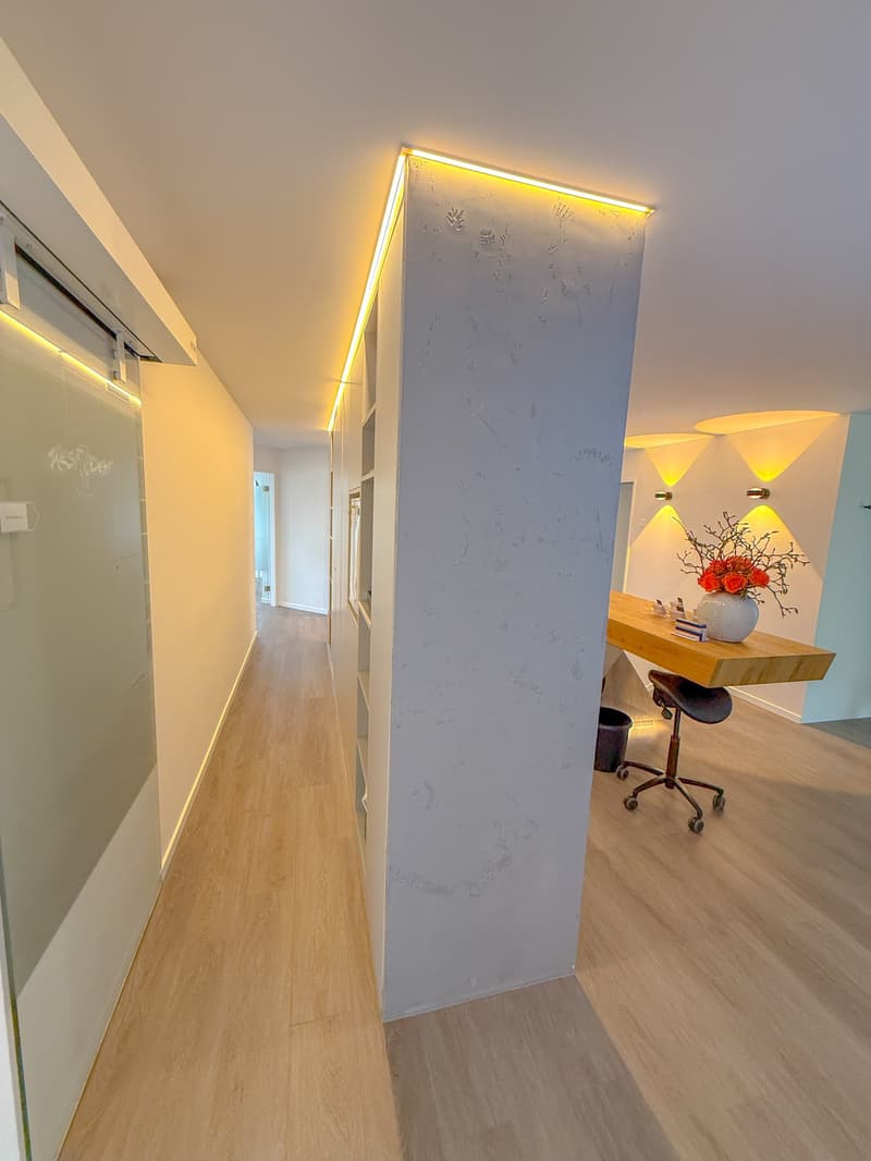 An empty hallway with wooden flooring and two lit up walls. A desk with a vase and a chair are on the right side. A door at the end of the hallway is open.