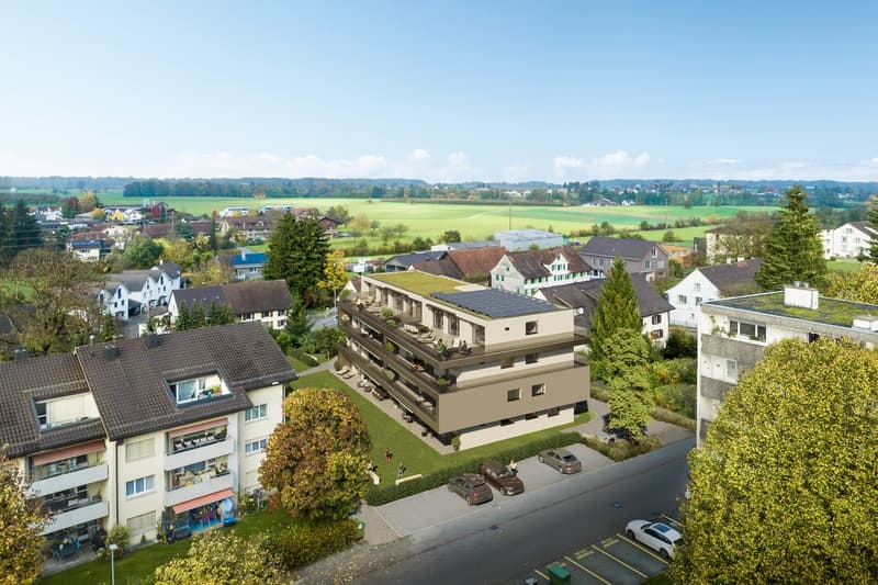 Multi-story residential building with balconies, surrounded by trees and greenery, with a parking lot in the foreground. The building has a modern, angular design with solar panels on the roof. The surrounding area appears to be a small town or village se