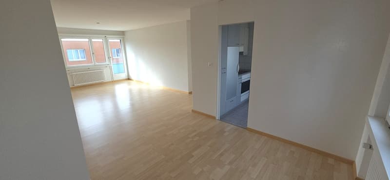 Empty room with wooden floors and white walls, an open door to the kitchen with a fridge, stove, and cabinets, a radiator below a window, and an open balcony.