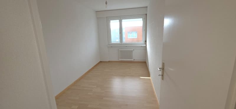 A spacious and empty room with wooden floors, white walls, and a window overlooking a building. A radiator is installed on the wall below the window.