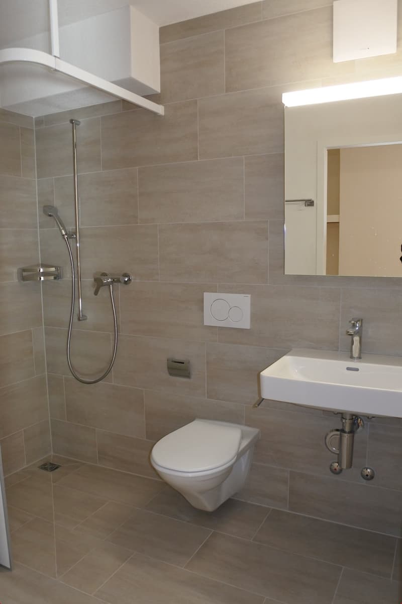 Bathroom with white toilet, white sink, mirror, shower head, and tiled walls