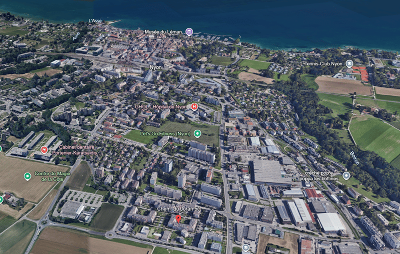 Aerial view of a city with various buildings, roads, and green spaces. Notable landmarks include the Musée du Léman, Tennis-Club Nyon, GHOL-Hôpital de Nyon, Let's Go Fitness (Nyon), and Centre de Magie de la Côte.