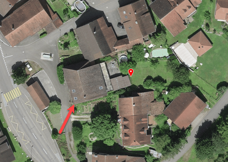 Aerial view of a residential area with several houses, driveways, and a road intersection. The houses have brown roofs and are surrounded by green grass and trees.