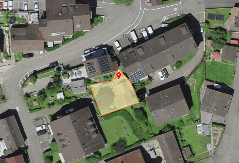 Aerial view of a residential area with several houses, a parking lot, and a road. The houses are brown with solar panels on the roofs. The parking lot has several cars parked. The road is surrounded by grass and trees. The area is well-lit, and the shadow