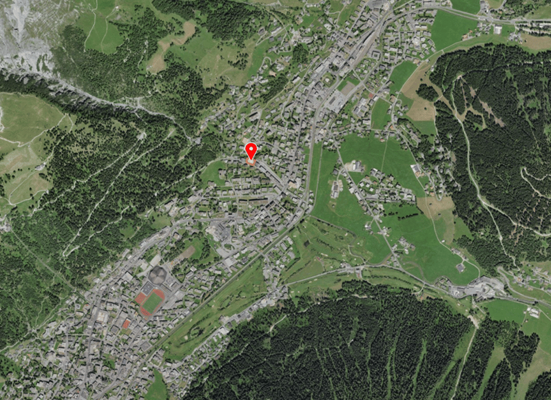 Aerial view of a town with many buildings, surrounded by green fields and trees, with a red pin indicating a specific location.