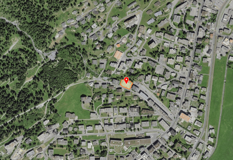 Aerial view of a town with many buildings, surrounded by green fields and trees, with a red marker indicating a specific location.