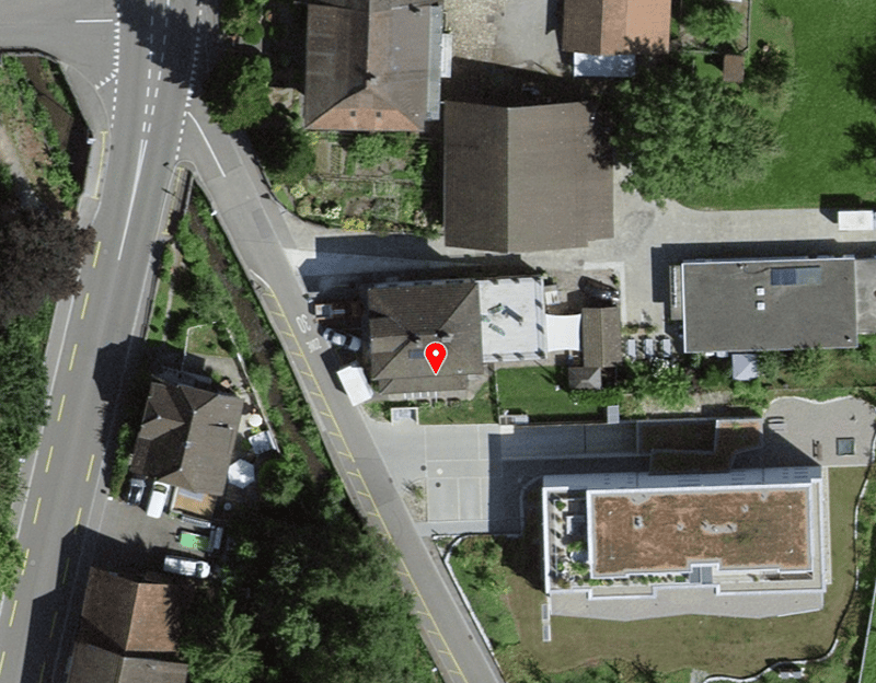 Aerial view of a neighborhood with several houses and a road. The houses are of different sizes and styles, with some having gardens and others having driveways. The road is a main street with a yellow line and a white line, and there are cars parked on t