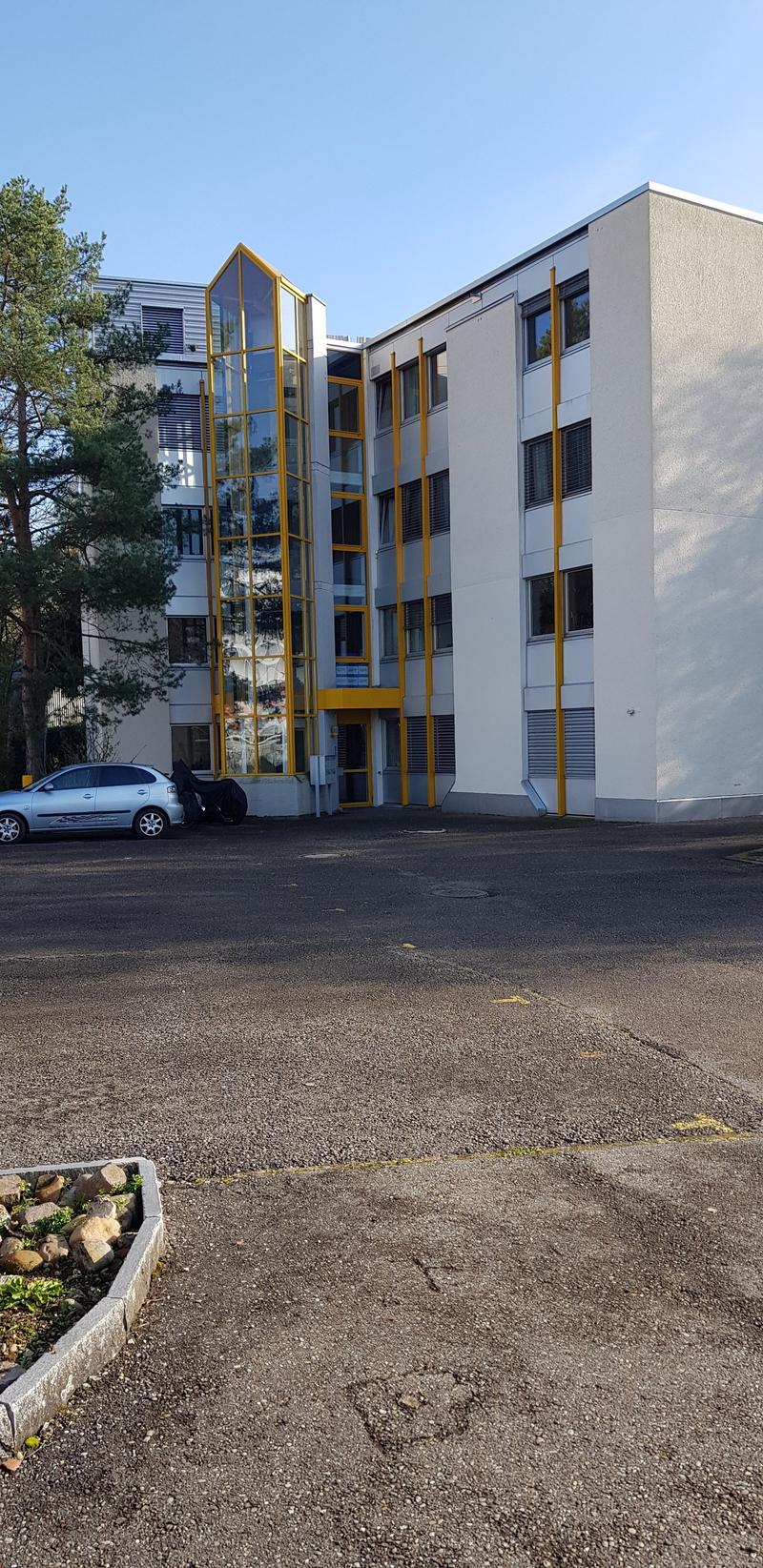 A multi-story building with glass and yellow accents. A parking area in front of the building with a parked car and some greenery.