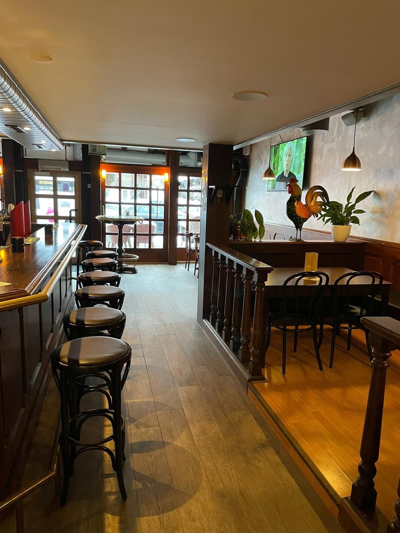 Seating area with bar stools, wooden chairs, and tables; a counter with a monitor; wooden floor, glass walls with doors, potted plants, and a decorative statue.